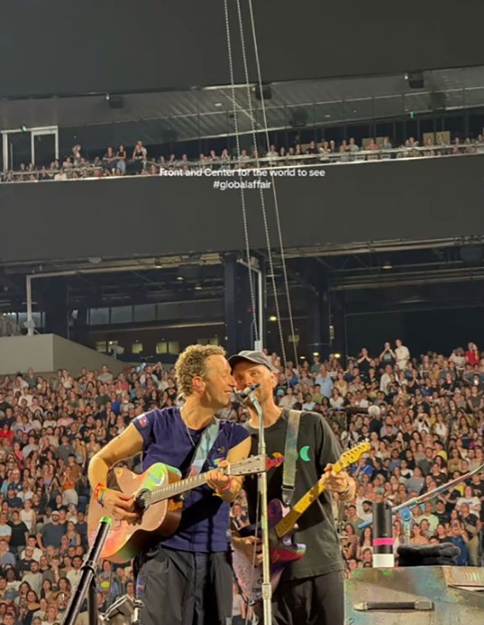 Coldplay band performing live on stage with guitars in front of a large excited concert audience.