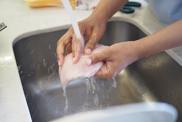 Person washing chicken under running water in a kitchen sink as part of cooking preparation and food safety discussion.