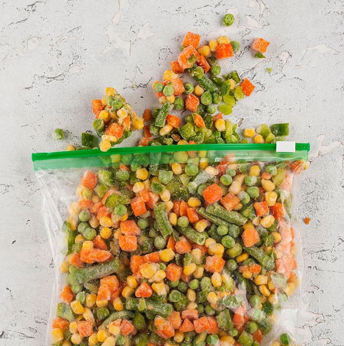 Frozen mixed vegetables spilling out of a resealable plastic bag on a textured light surface.