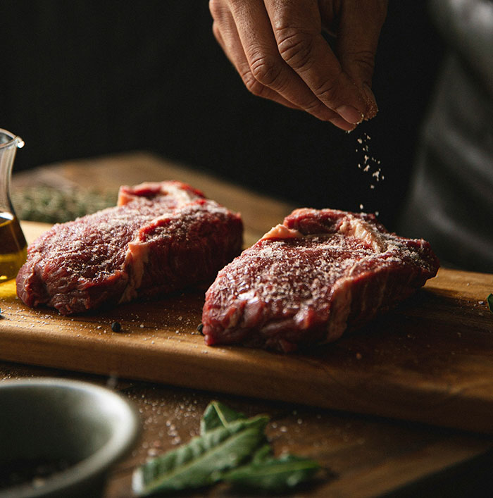 Close-up of raw steaks being seasoned on a wooden cutting board, illustrating cooking preparation and seasoning techniques.