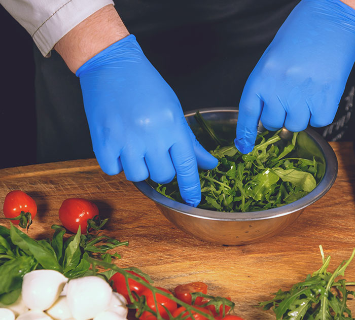 Hands wearing blue gloves mixing greens in a metal bowl on wooden surface with fresh vegetables nearby, washing chicken myth.
