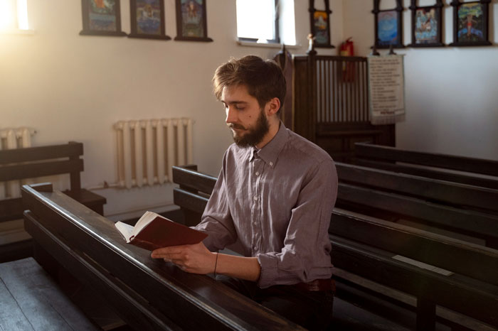 Man sitting in church pew reading a book, representing religious beliefs linked to feminine hygiene waste bins issue.