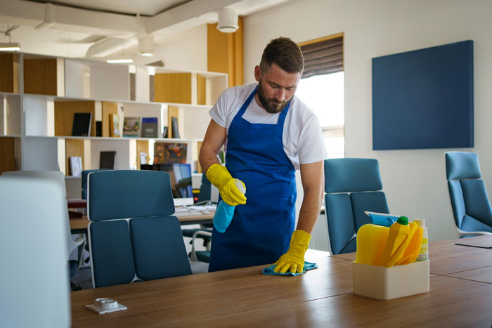Custodian cleaning office, wearing gloves and apron, handling waste bins with challenges related to religion.
