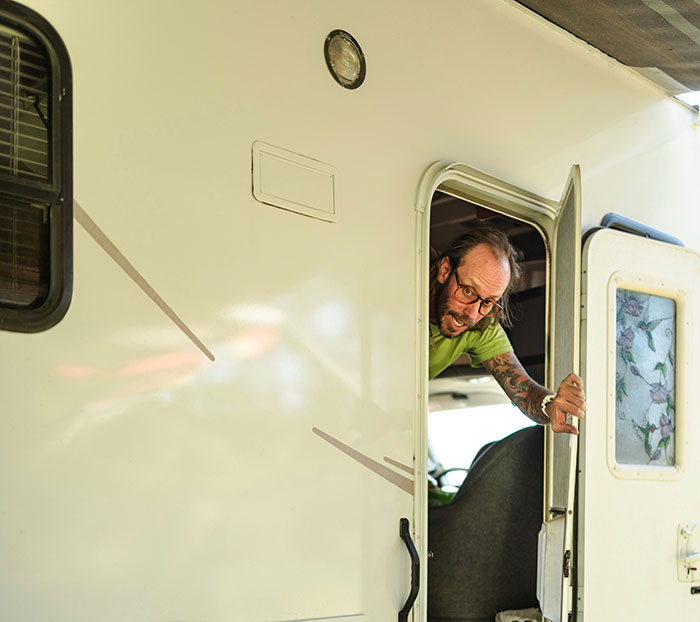 Man with glasses and tattoos leaning out of a camper door, surprising medical professionals with his survival story.