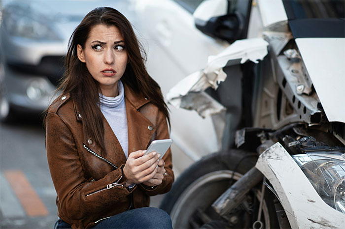 Woman in brown jacket holding phone, looking scared beside a damaged white car, depicting creepy and terrifying facts scenario.