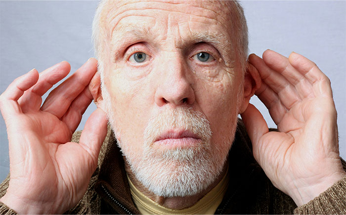 Elderly man with a serious expression using hands to cup ears, symbolizing awareness of creepy and terrifying facts.