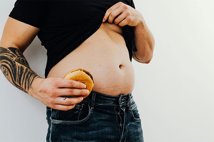 Man lifting shirt to reveal belly while holding a burger, illustrating creepy and terrifying facts about health risks.
