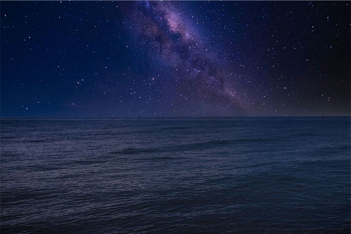 Starry night sky over a dark ocean conveying eerie and creepy atmosphere with distant faint lights on the horizon.