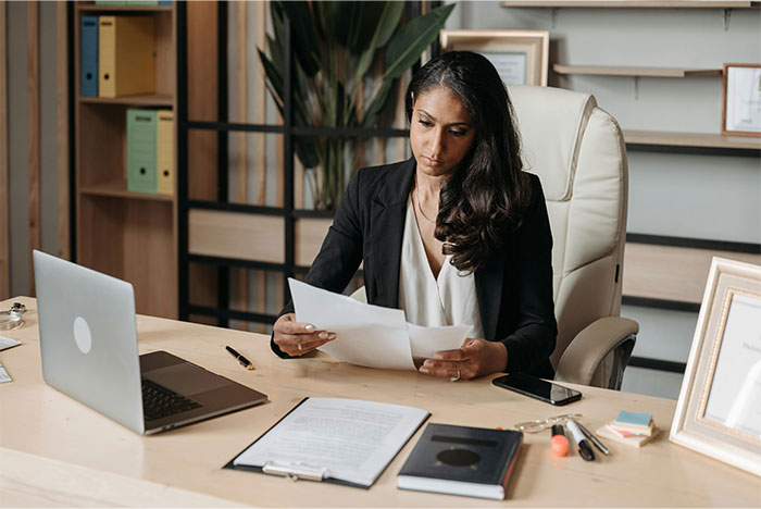 Woman in black blazer reading papers at desk with laptop, phone, and office supplies, reflecting on creepy and terrifying facts.