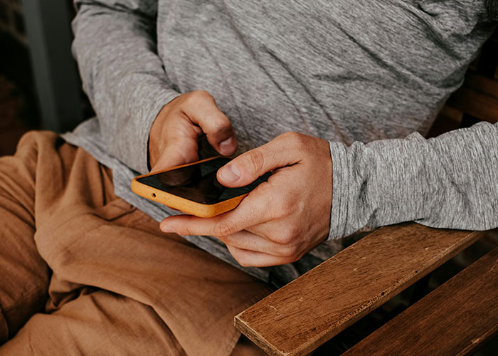Man in gray shirt and brown pants holding a smartphone, representing a dad concerned about 8-year-old spending the night.
