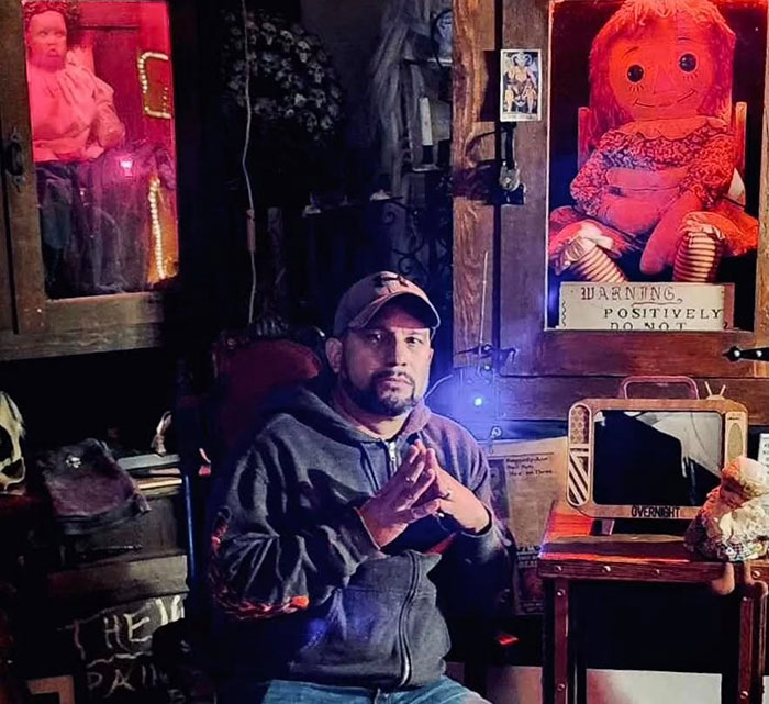 Paranormal investigator sitting in a dim room with the possessed Annabelle doll displayed behind him on a wooden shelf. - 1