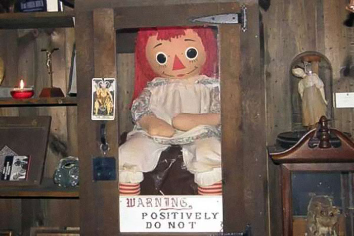 Possessed Annabelle doll displayed in a wooden case with warning sign inside a dimly lit paranormal investigation room. - 3