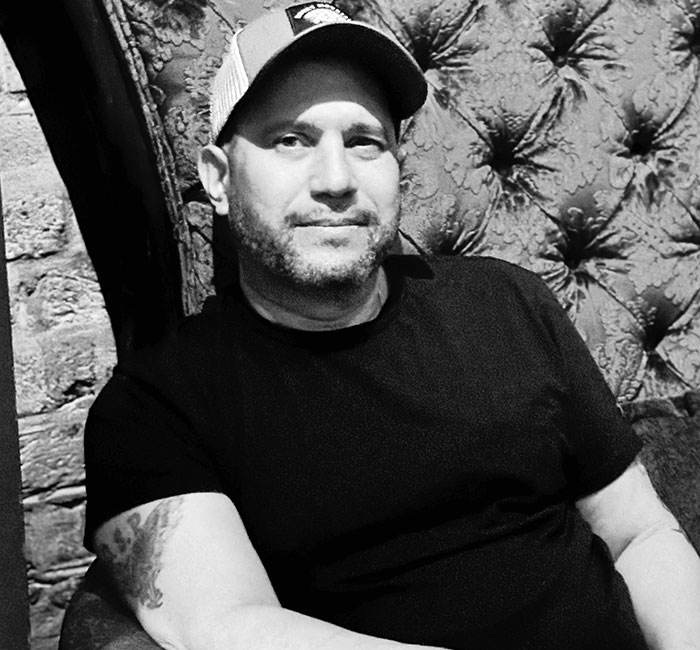 Man wearing a baseball cap and black shirt, sitting against a patterned wall, related to paranormal investigator news. - 5