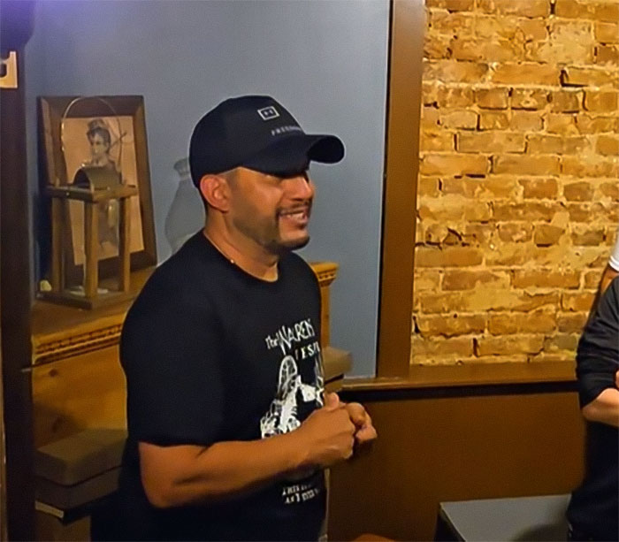 Man wearing a black cap and paranormal investigator shirt speaking during a tour with possessed Annabelle doll in a dim room. - 7