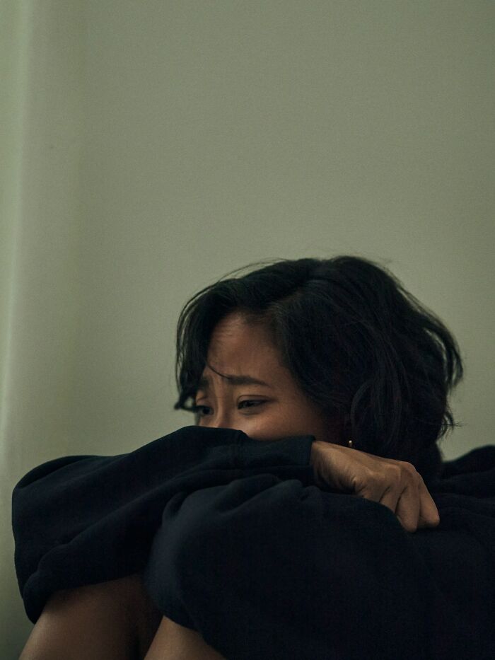 Woman with dark hair and black sweater sitting against wall, showing signs of distress and mysterious body quirks.