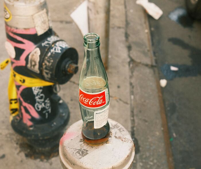 Half-empty Coca-Cola bottle on a dirty public street next to a graffiti-covered fire hydrant and caution tape.