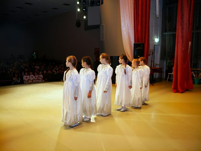 Six girls in white dresses stand in a line on stage facing sideways during a performance in front of an audience.