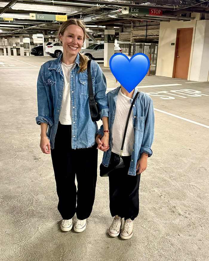 Kristen Bell smiling in a parking garage wearing denim jacket and holding hands with a child in similar outfit.