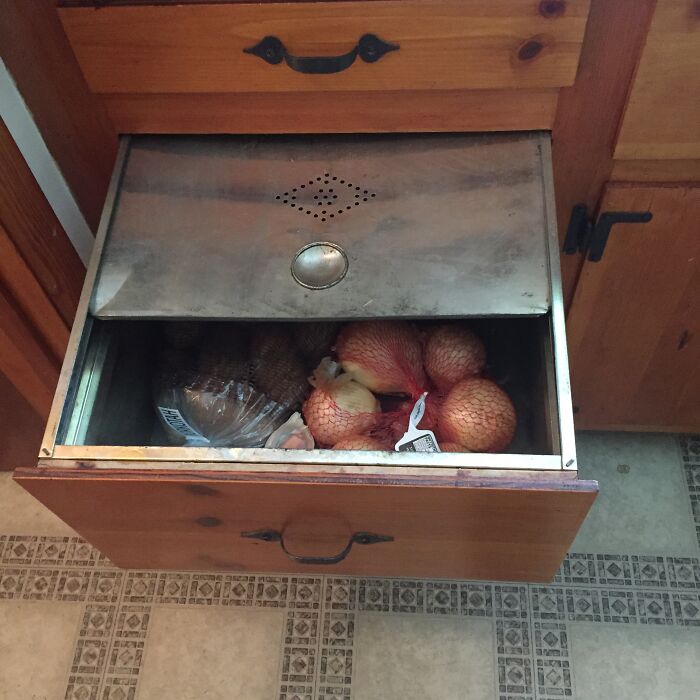 1950s kitchen vegetable storage drawer with metal vent, wooden cabinets, and mesh bags of onions inside.