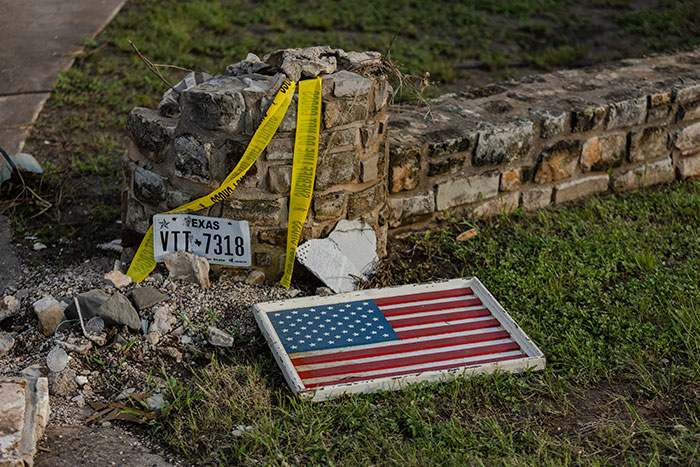 Damaged stone wall with Texas license plate and fallen American flag, marked with caution tape after flood disaster.