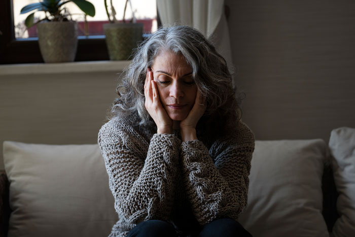 Woman with gray hair sitting on a couch, looking thoughtful and holding her face, representing delivery room baby DNA concerns. - 4