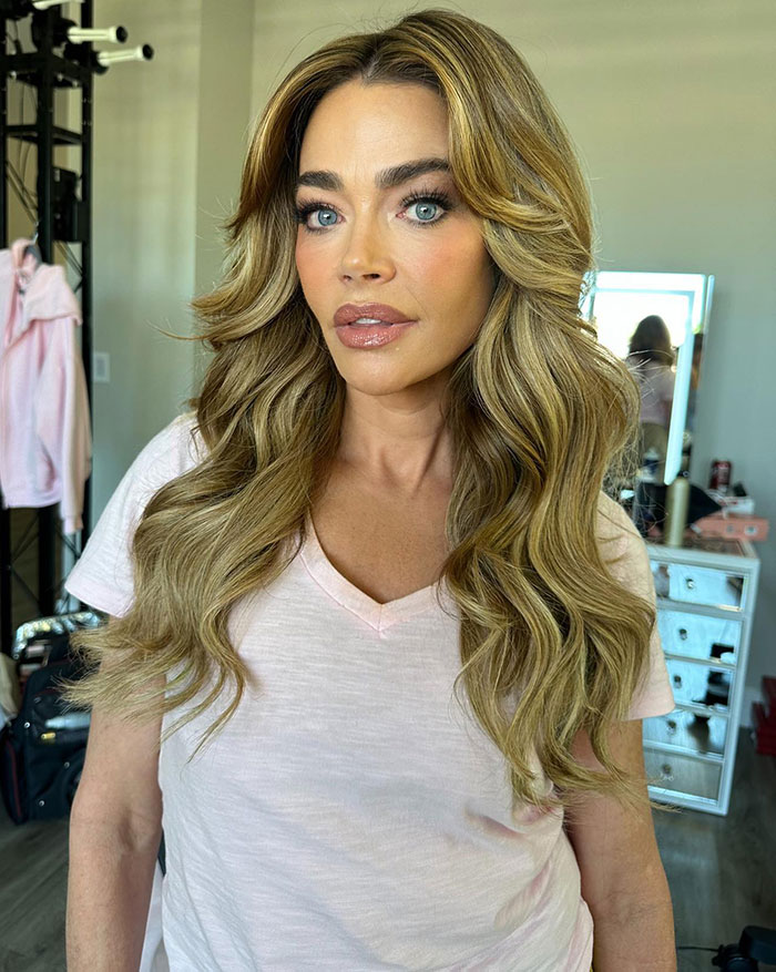 Denise Richards with styled blonde hair and makeup, posing indoors amid personal items and a mirror in the background Denise Richards with styled blonde hair and makeup, posing indoors amid personal items and a mirror in the background