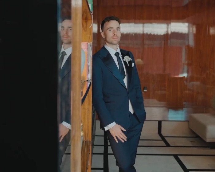 Man in a formal navy suit standing indoors, reflecting on life's fragility with a solemn and thoughtful expression.
