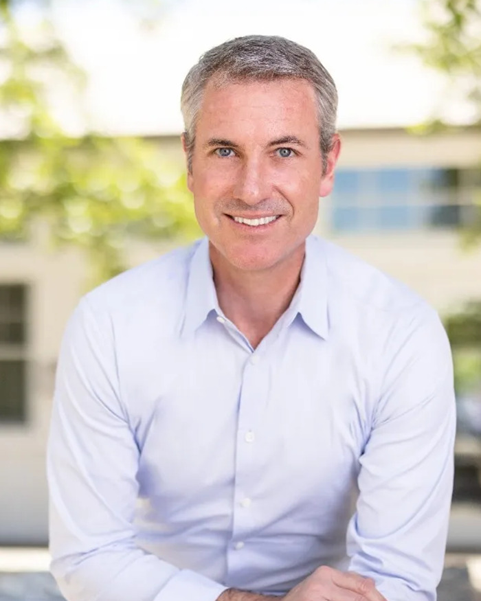 Disgraced CEO Andy Byron smiling in a light blue shirt, posing outdoors with blurred greenery and building in the background