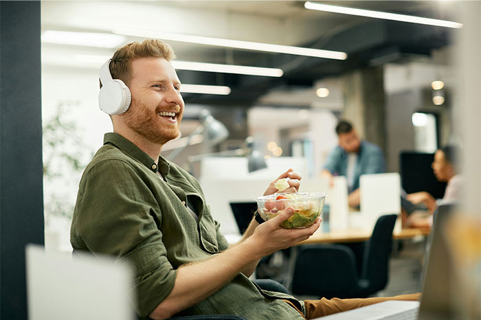 Man wearing headphones eating salad and laughing in a modern office, capturing a not my proudest moment feeling. - 42
