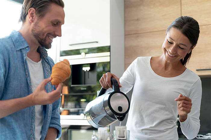 A smiling woman pours coffee from a kettle while a man in a denim shirt holds a croissant in a modern kitchen. - 5