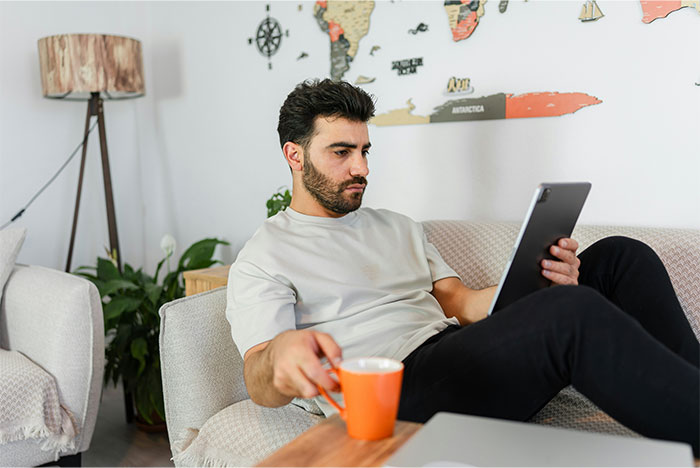 Man relaxing on sofa, using tablet with one hand and holding an orange mug, showing a moment of stopping helping others. - 18