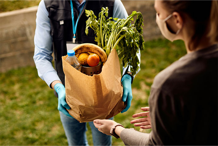 Person wearing gloves and mask handing a paper bag of groceries to another person outdoors, illustrating stopped helping others concept. - 50