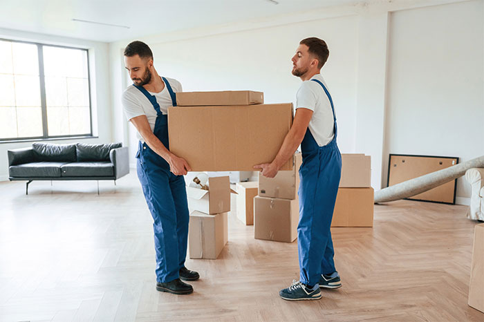 Two movers in blue overalls carrying a large cardboard box inside a modern apartment with packed boxes around. - 31