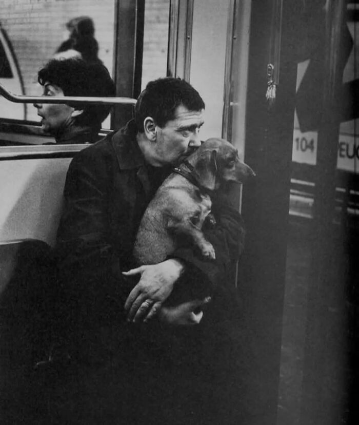 Man sitting on a bus holding and kissing his dog, a random animal sighting captured on the street in black and white.