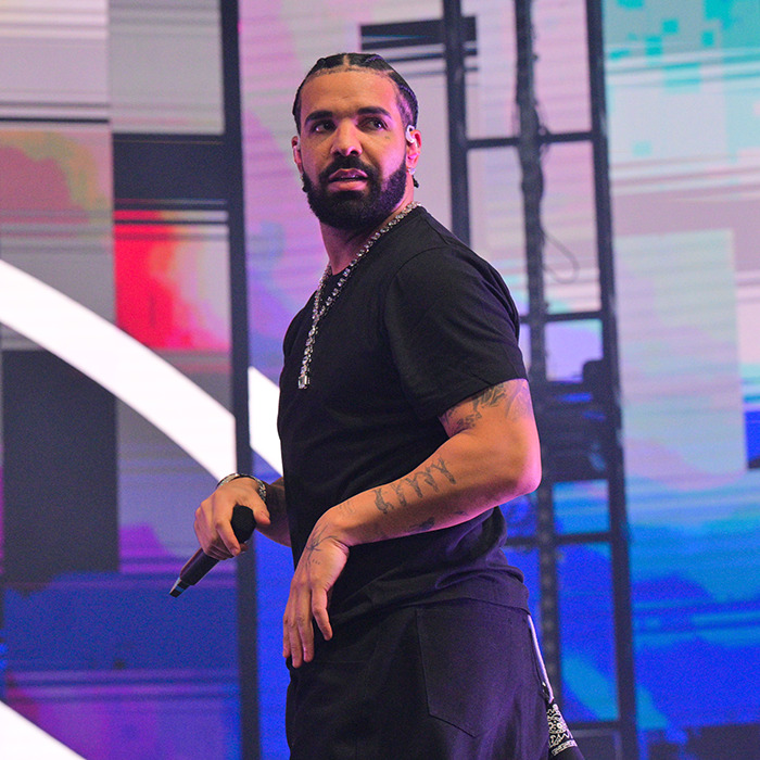 Drake on stage holding a microphone, with visible tattoos and casual black outfit sparking plastic surgery for his abs rumors.
