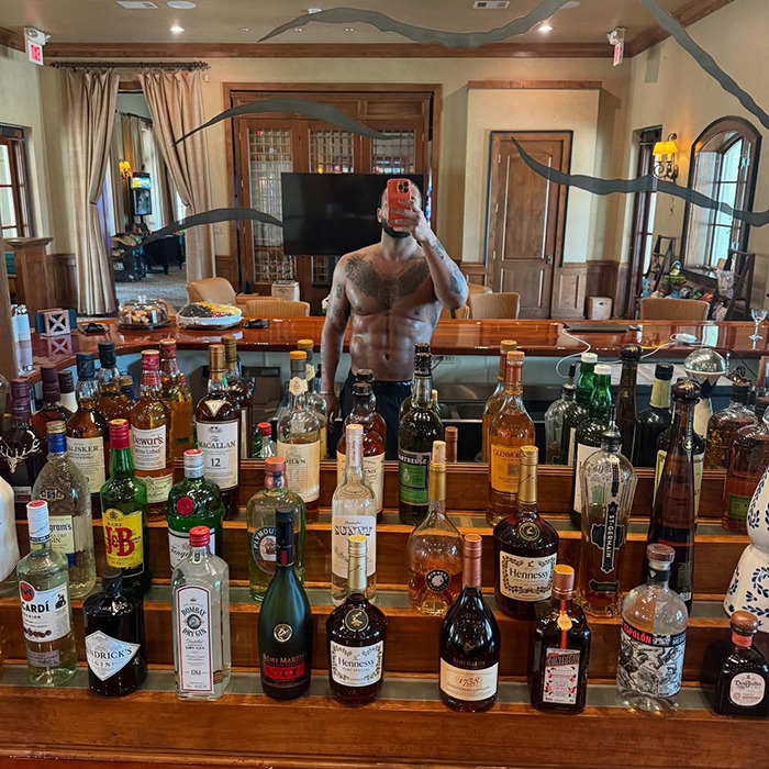 Shirtless man with visible abs taking a mirror selfie behind a bar filled with assorted liquor bottles indoors.
