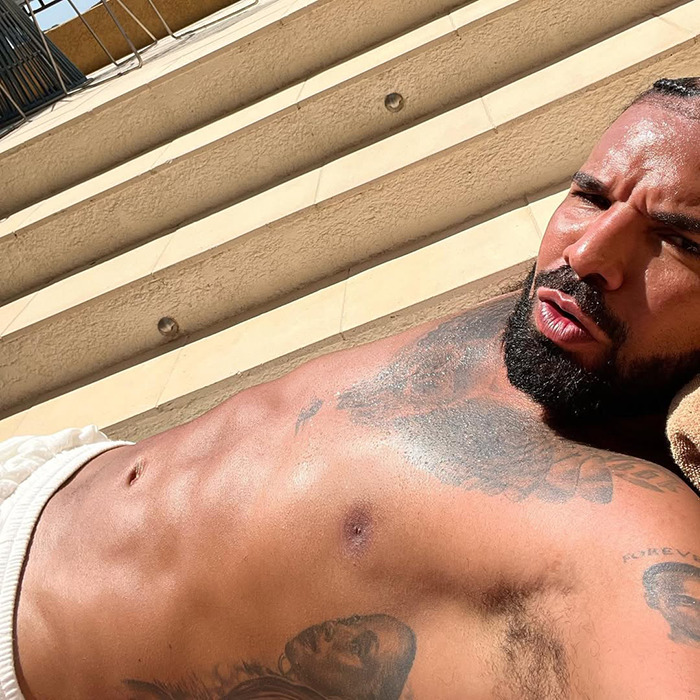 Drake shirtless showing his abs and tattoos while relaxing in the sun, sparking plastic surgery and fake six-pack rumors.
