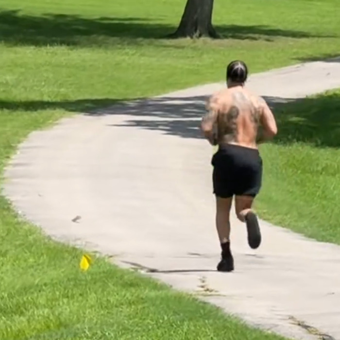 Shirtless man jogging on a curved path in the park, showing back muscles and tattoos, raising questions about plastic surgery abs.