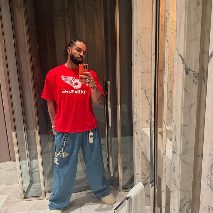 Man in red shirt and loose pants taking a mirror selfie, related to plastic surgery and fake six-pack abs debate.