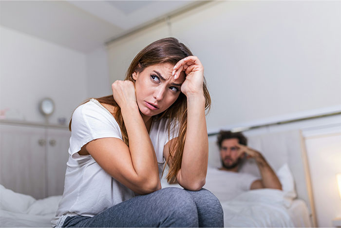 Woman sitting on bed looking distressed while man watches from behind, depicting tension and conflict in a relationship. - 18
