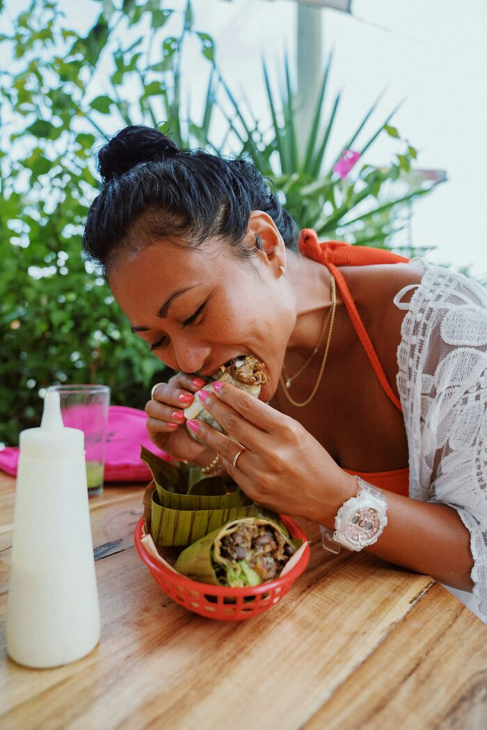 Woman with body quirks eating wrapped food outdoors at a wooden table with greenery in the background.