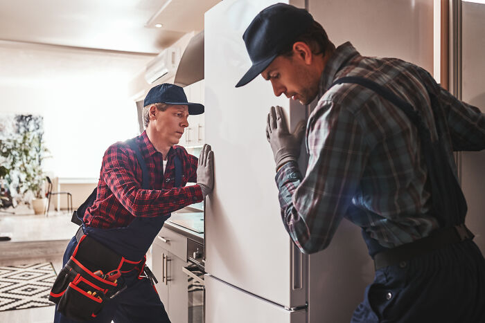 Two workers wearing gloves and plaid shirts moving a refrigerator, illustrating things non-Americans do that confuse Americans. - 8