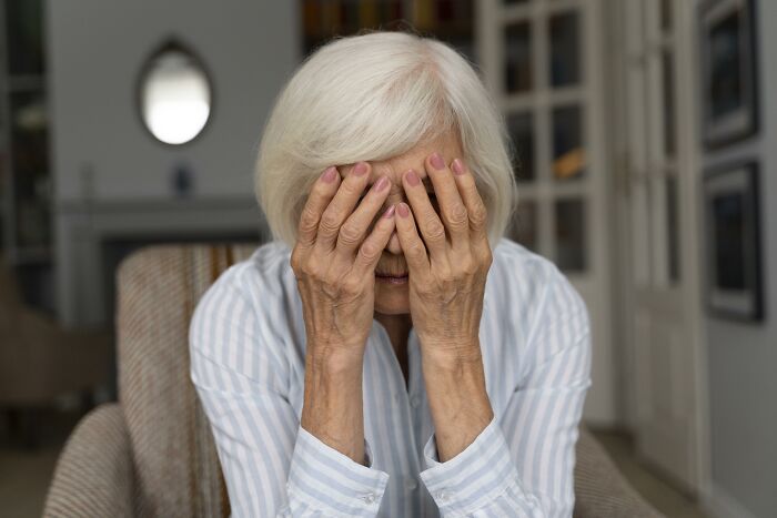 Elderly woman covering her face with hands, reflecting on unhinged pieces of family lore in a quiet home setting.