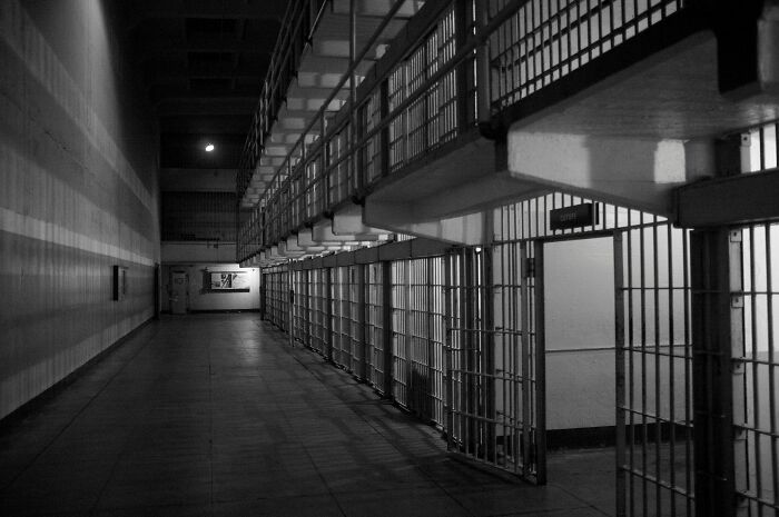 Black and white image of a dimly lit prison corridor with cells and metal bars along both sides.
