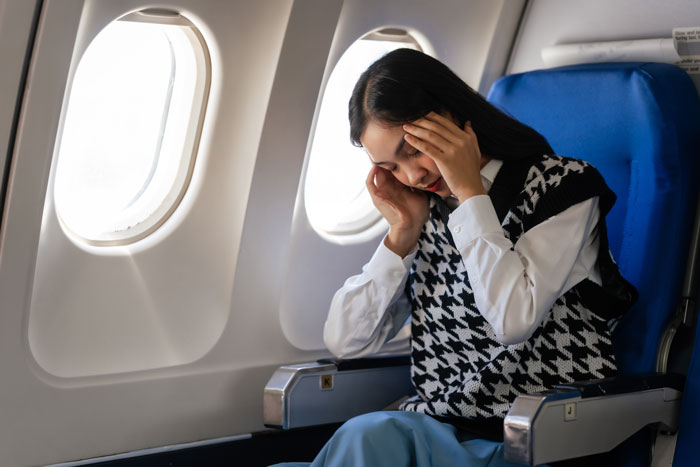 Young woman sitting on a plane seat looking stressed, illustrating a cancer survivor moved due to entitled passenger’s child. Young woman sitting on a plane seat looking stressed, illustrating a cancer survivor moved due to entitled passenger’s child.
