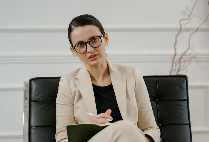 Woman in beige suit and glasses sitting on black chair, holding pen and notebook, appearing concerned during discussion. Woman in beige suit and glasses sitting on black chair, holding pen and notebook, appearing concerned during discussion.
