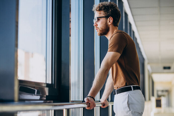 Young escape room employee wearing glasses, looking thoughtfully out the window during a break in the hallway. - 26