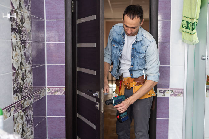 Escape room employee in casual outfit using a drill to fix a door in a room with purple floral tiles. - 22