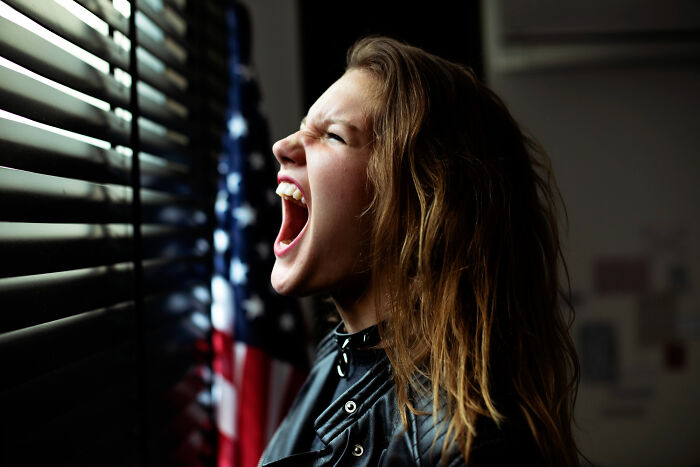 Young woman with long hair shouting by window blinds next to an American flag, relating to escape room employees stories. - 15