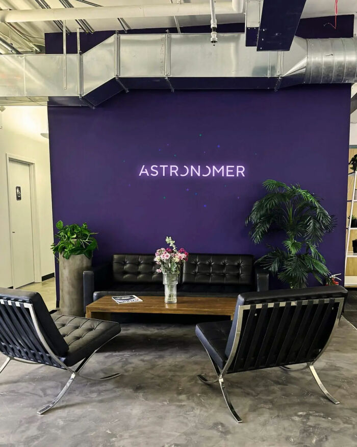 Modern office lounge with Astronomer logo, black leather seating, plants, and a wooden coffee table, reflecting toxic CEO drama.